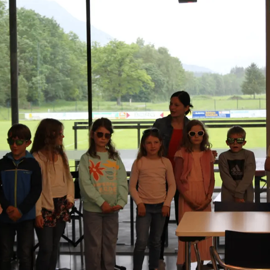 Eine Gruppe von Kindern steht zusammen mit einer Frau in einem Raum mit großen Fenstern, die auf ein Feld und Berge blicken. Sie tragen alle Sonnenbrillen.