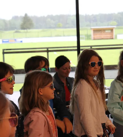 Eine Gruppe junger Mädchen mit Sonnenbrillen steht in der Nähe einer Glaswand, wobei ein Feld und ein Gebäude im Hintergrund sichtbar sind.
