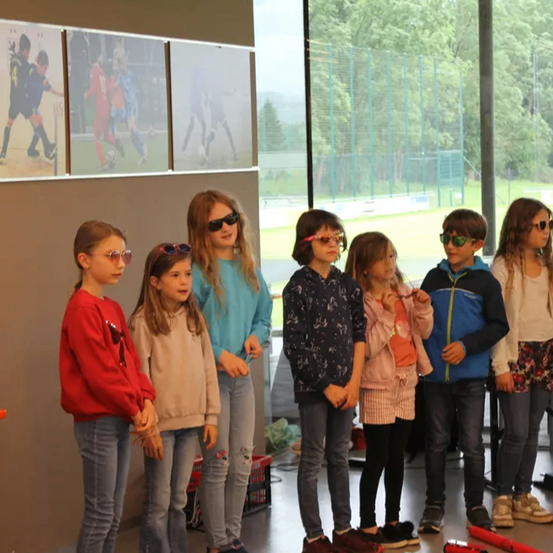 Eine Gruppe von Kindern mit Sonnenbrillen steht vor einer Wand mit Fußballbildern, draußen ist ein Feld zu sehen.