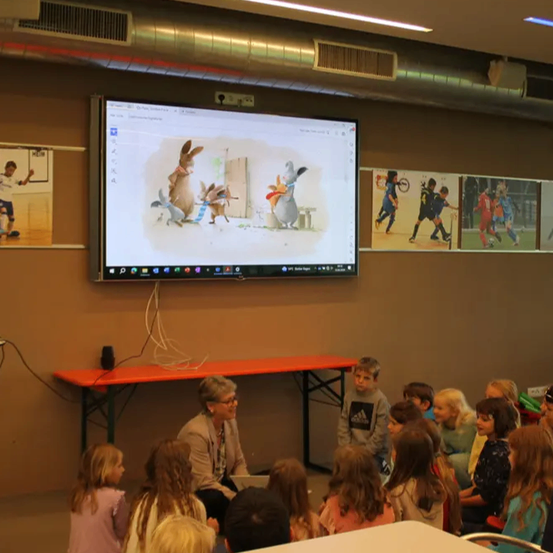 Eine Frau sitzt an einem roten Tisch mit einem Laptop vor einem Bildschirm, der ein animiertes Bild von Kaninchen zeigt. Kinder sitzen vor ihr und schauen zu.