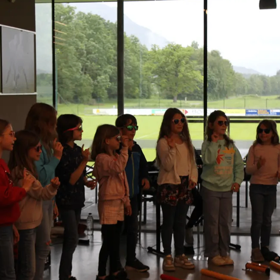 Eine Gruppe von Kindern, alle mit Sonnenbrillen, steht in einer Reihe in einem Raum mit großen Glasfenstern. Sie schauen auf ein Feld mit Bäumen und Bergen in der Ferne.