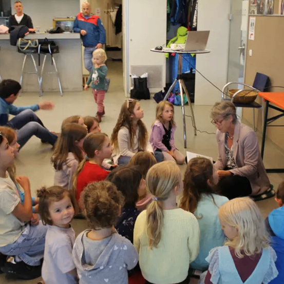Eine Gruppe junger Mädchen und Jungen sitzt auf dem Boden in einem Raum mit einer Frau vor ihnen. Einige Erwachsene sitzen und stehen im Hintergrund. Ein Laptop steht auf einem Tisch.