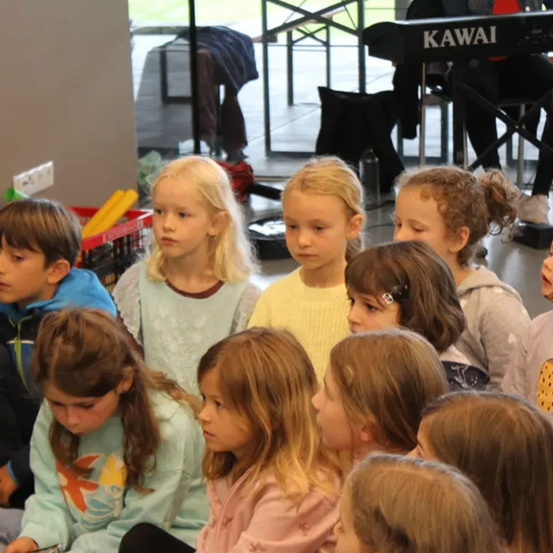Eine Gruppe junger Kinder, Mädchen und Jungen, sitzt in einem Kreis, möglicherweise für eine Schulveranstaltung. Sie alle schauen in die gleiche Richtung. Im Hintergrund steht ein Kawai-Piano.