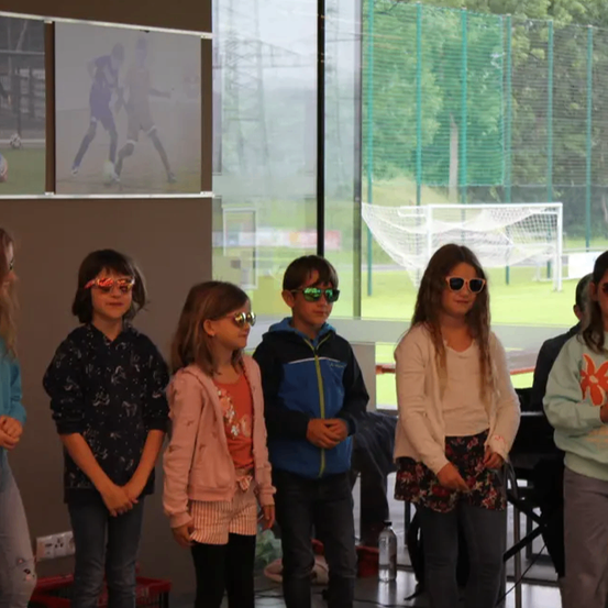 Eine Gruppe junger Kinder in Sonnenbrillen steht zusammen. Sie tragen Freizeitkleidung und scheinen für ein Foto zu posieren. Hinter ihnen befindet sich eine Wand mit Bildern von Fußballspielern.