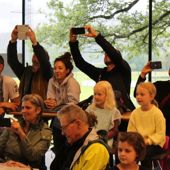 Eine Gruppe von Menschen macht Fotos in einem Glasgebäude mit Bäumen draußen. Einige sitzen, andere stehen und alle halten eine Kamera.