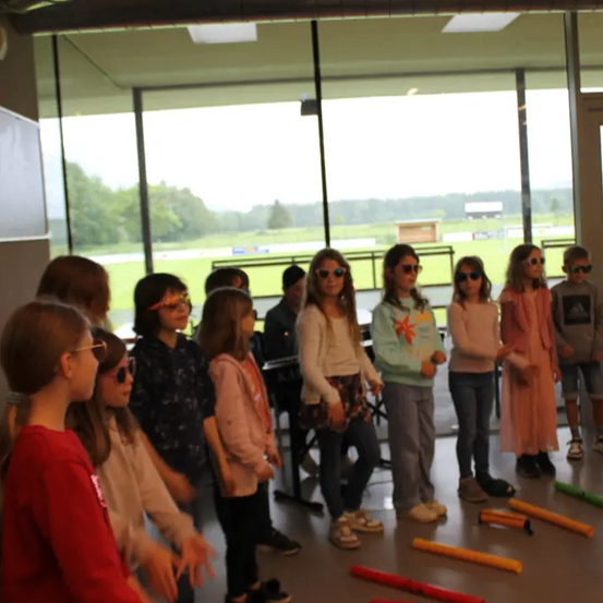 Eine Gruppe von Kindern steht in einer Reihe in einem Klassenzimmer, trägt Sonnenbrillen und hält Musikinstrumente. Das Klassenzimmer hat Glaswände und draußen ist ein Feld.