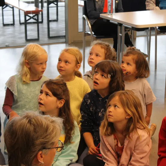 Eine Gruppe junger Mädchen und eine erwachsene Frau sitzen und hören aufmerksam einem Sprecher zu. Sie befinden sich in einem Raum mit Stühlen, Tischen und einer Glaswand.