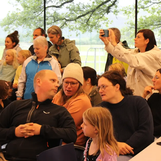 Eine Gruppe von Menschen, einige sitzend und andere stehend, versammelt sich drinnen. Eine Frau macht ein Foto mit ihrem Handy. Bäume und ein Grasfeld sind durch die Glaswände zu sehen.