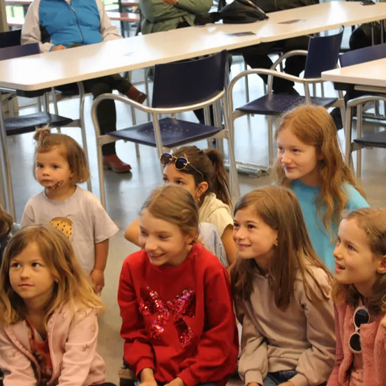 Eine Gruppe junger Mädchen sitzt auf Stühlen in einem Klassenzimmer. Einige von ihnen tragen Brillen und eine hat eine Sonnenbrille auf ihrem Pullover. Einige Erwachsene sitzen im Hintergrund an Tischen.
