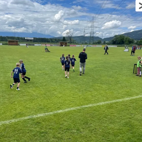 Eine Gruppe von Kindern spielt Fußball auf einem Feld unter einem teilweise bewölkten Himmel. Einige laufen, während andere stehen. Ein Mann geht in der Nähe.