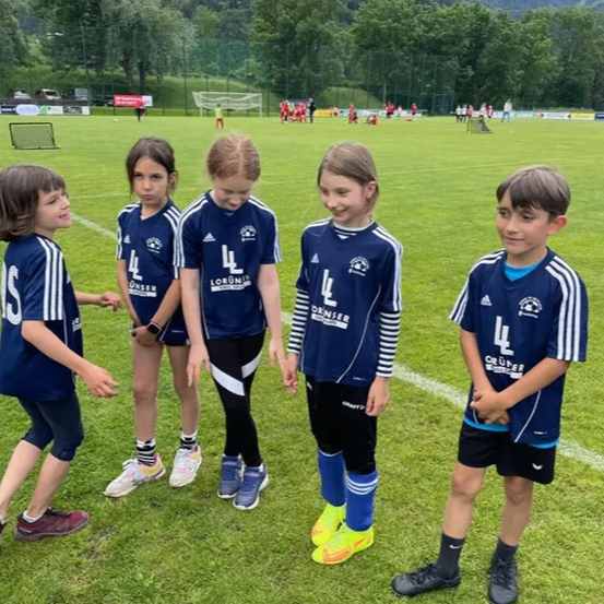 Fünf Kinder, möglicherweise Fußballspieler, stehen auf einem Fußballfeld zusammen und tragen blaue Uniformen mit weißen Streifen. Die Kinder schauen auf etwas vor ihnen. In der Ferne befinden sich Berge und Bäume.