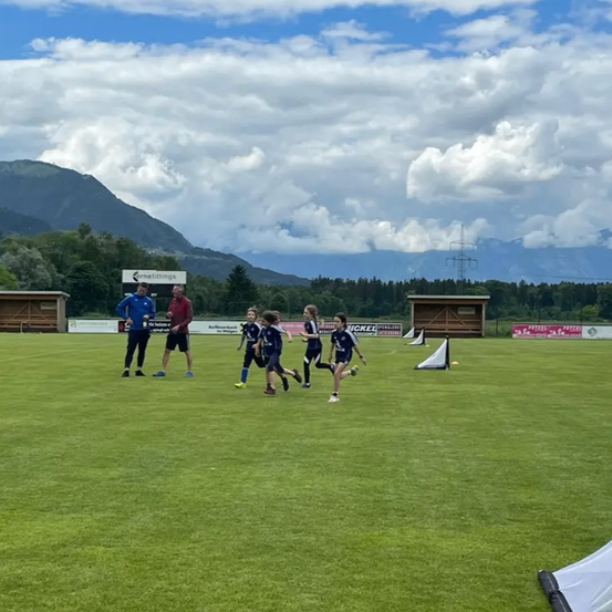 Eine Gruppe von Kindern läuft auf einem Feld unter einem bewölkten Himmel, mit Bergen im Hintergrund. Zwei Erwachsene stehen und beobachten sie.