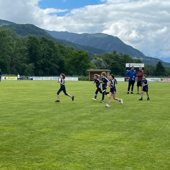Eine Gruppe von Kindern läuft auf einem Feld mit Bergen im Hintergrund. Es stehen zwei Männer in der Nähe, einer trägt eine blaue Jacke.