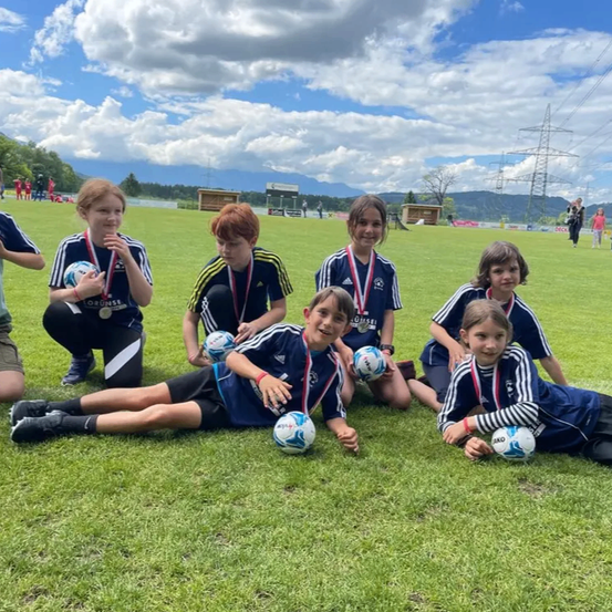 Eine Gruppe junger Fußballspieler posiert mit ihren Medaillen an einem sonnigen Tag, mit Bergen und Stromleitungen im Hintergrund.