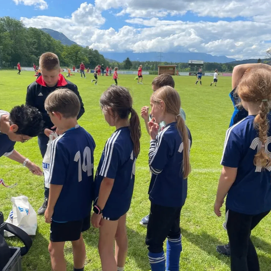 Eine Gruppe von Kindern in blauen Trikots steht auf einem Rasenfeld. Ein Erwachsener mit Brille spricht mit ihnen. Im Hintergrund spielen andere Kinder Fußball.