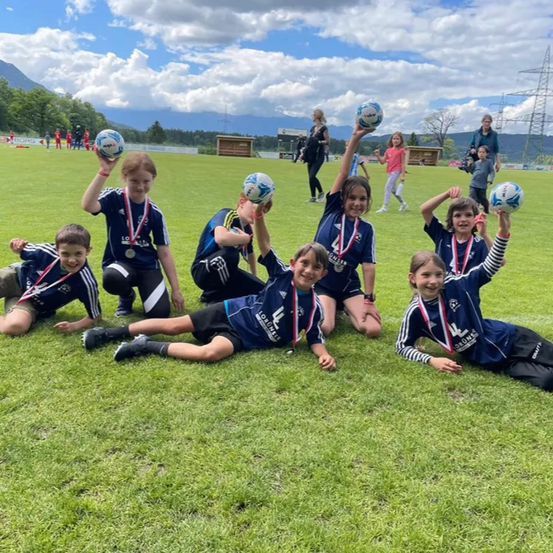 Eine Gruppe junger Athleten in blauen Trikots posiert auf einem Rasenfeld und hält Fußballbälle und Medaillen. Dahinter stehen eine Frau und ein Kind auf dem Feld.