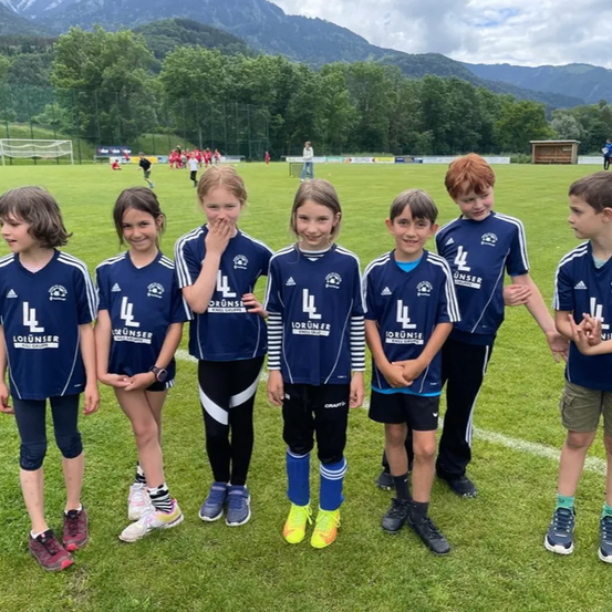 Sieben Kinder in blau-weißen Uniformen stehen in einer Reihe auf einem Fußballfeld. Sie lächeln und posieren für ein Foto. Im Hintergrund sind ein Bergrücken und ein Fußballtor zu sehen.