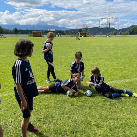 Fünf Kinder in Sportuniformen sind auf einem Rasenfeld. Vier sitzen oder liegen und halten Fußballbälle. Eins steht mit einem Ball. Berge und Bäume sind im Hintergrund.