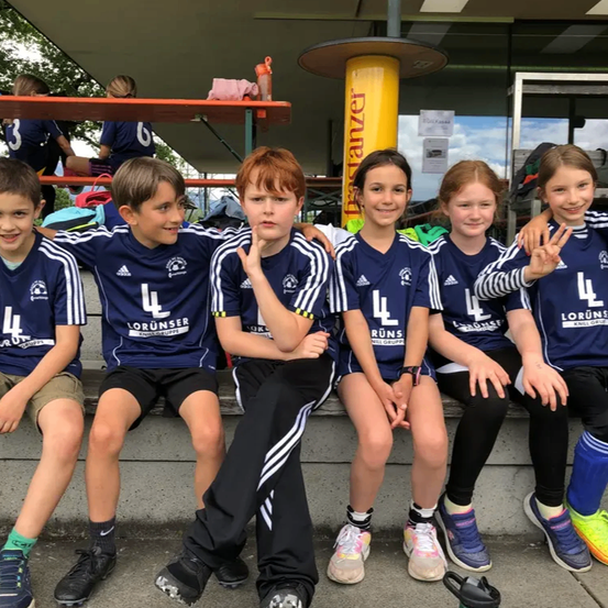 Sechs Kinder in passenden blauen Trikots mit dem Logo 'LorUnser' sitzen auf einer Betonbank, lächeln und posieren für ein Foto. Im Hintergrund ist ein Gebäude mit Glasfenstern und einer Metallstruktur zu sehen.