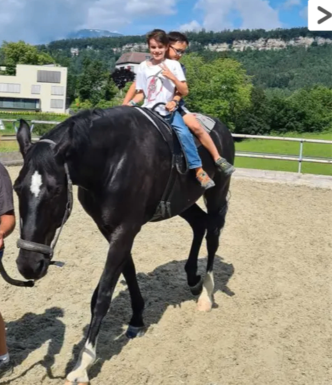 Zwei Kinder reiten auf einem schwarzen Pferd in einem Außenbereich. Hinter ihnen ist ein Zaun, und es gibt Bäume und ein Gebäude.