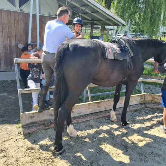 Ein Mann in einem blauen Hemd hilft einem jungen Mädchen, ein schwarzes Pferd in einem Stall zu besteigen, während andere in der Nähe sind.