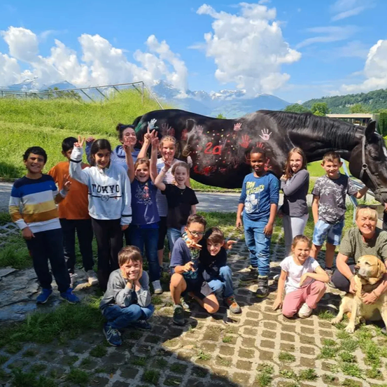 Eine Gruppe von Kindern und zwei Erwachsenen posiert für ein Foto mit einem Pferd und einem Hund. Das Pferd hat Handabdrücke und die Nummer 24 auf seinem Körper. Sie befinden sich in einem Grasbereich mit Bergen im Hintergrund.
