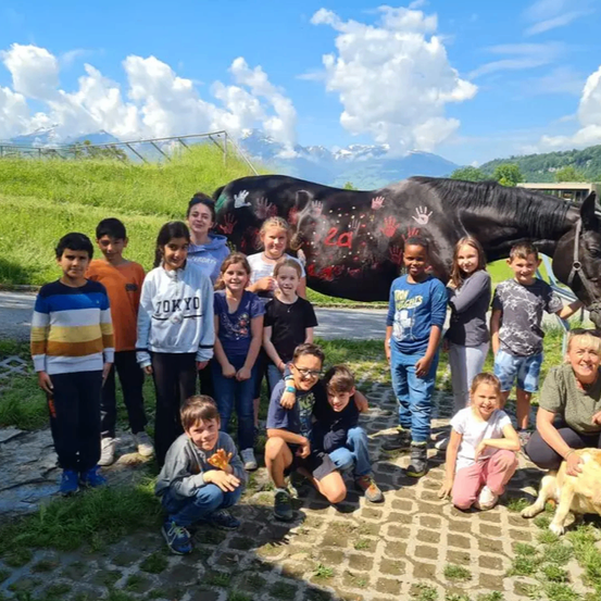 Eine Gruppe von Kindern und einem Erwachsenen posiert für ein Foto mit einem Pferd, dessen Körper mit gemalten Händen verziert ist, in einem Grasbereich.