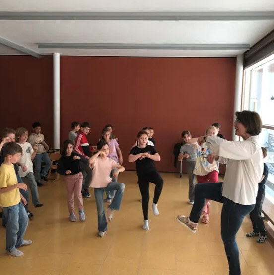 Eine Gruppe von Kindern und einem Erwachsenen übt Tanzbewegungen in einem Raum mit roten Wänden und Holzböden. Der Erwachsene leitet die Übung.