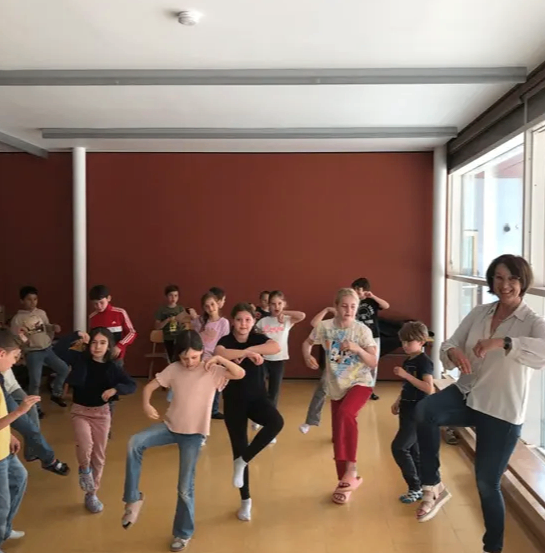 Eine Gruppe von Kindern und eine erwachsene Frau üben Tanzschritte in einem Raum mit roten Wänden und großen Fenstern. Die Kinder tragen Turnschuhe und die Frau ist in Sandalen.