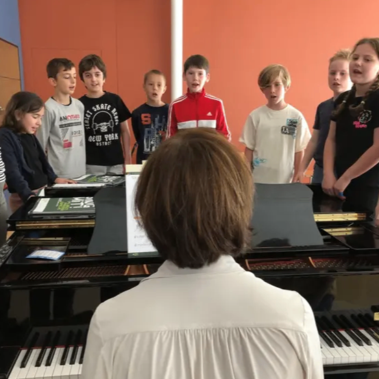 Eine Gruppe von Kindern, sowohl Jungen als auch Mädchen, singt, während sie hinter einem Klavier stehen, das von einer Frau gespielt wird.