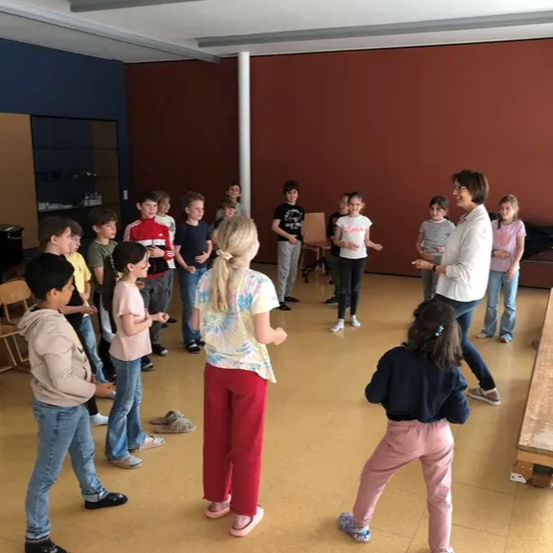 Eine Gruppe von Kindern steht in einem Kreis mit einer Frau, wahrscheinlich in einem Klassenzimmer. Sie schauen auf die Frau, die wahrscheinlich Anweisungen gibt. Der Raum hat geflieste Böden und braune Wände.