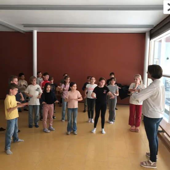 Eine Gruppe von Kindern und einem Erwachsenen in einem Klassenzimmer übt Tanzbewegungen, während sie einander ansehen.