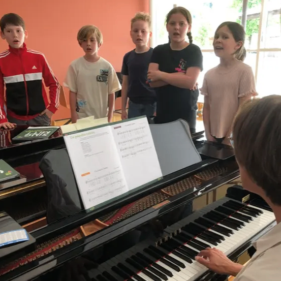 Eine Gruppe von Kindern beobachtet eine Person beim Klavierspielen, mit offenen Notenblättern vor ihnen.