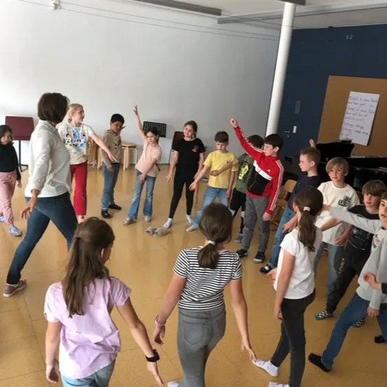 Eine Gruppe von Kindern und einer Lehrerin tanzen in einem Raum mit Holzböden. Die Kinder tragen verschiedene Kleidungsstücke und die Lehrerin trägt eine weiße Kapuzenjacke. Im Raum stehen Musikinstrumente.