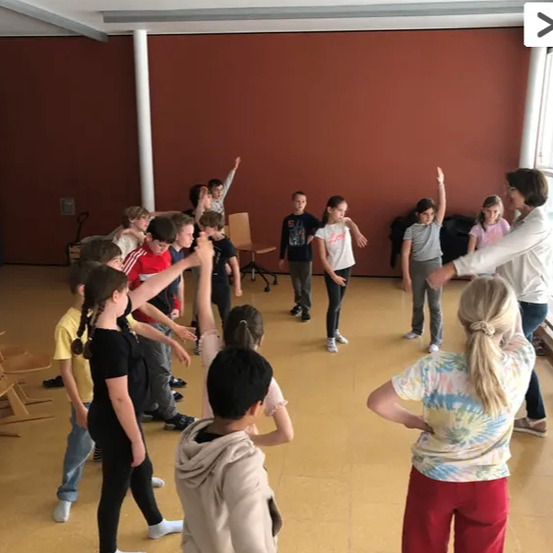 In einem Raum sind mehrere Kinder in einem Tanzkurs, angeführt von einer Frau. Der Raum hat Holzböden und rote Wände.