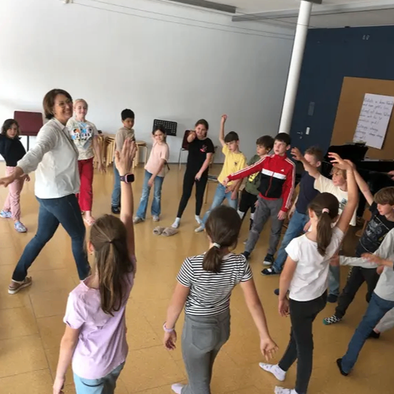 Eine Gruppe von Kindern in einem Tanzstudio, geleitet von einer erwachsenen Frau, die sie in Tanzschritten anleitet, während sie ihren Anweisungen folgen.