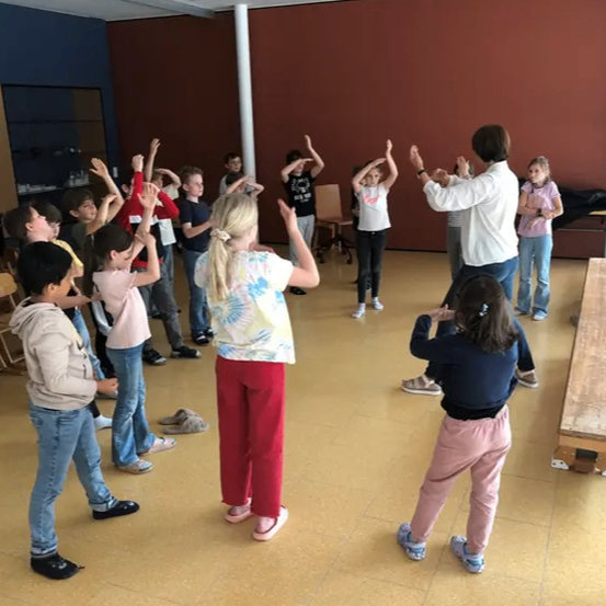 In einem Raum führen Kinder und ein Erwachsener Tanzbewegungen durch. Einige haben Namensschilder auf ihren Shirts, und eine Holztafel steht in der Ecke.