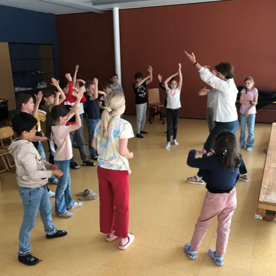 Eine Gruppe von Kindern und einem Erwachsenen in einem Raum nehmen an einer Tanzaktivität teil, wobei einige Kinder ihre Hände heben und andere auf dem gefliesten Boden tanzen.