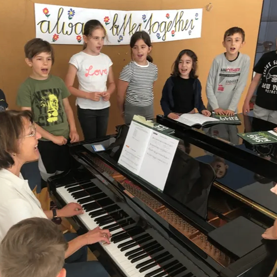 Eine Frau spielt Klavier, während mehrere Kinder vor ihr singen. Dahinter hängt ein Banner mit den Worten 'always ballet together' an der Wand.