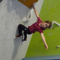 Ein junges Mädchen klettert an einer Kletterwand, mit ausgestreckten Armen und Beinen, in einem roten Shirt und schwarzen Hosen.
