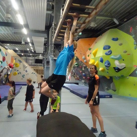 Ein junges Mädchen klettert eine Holzleiter in einem Fitnessstudio mit bunten Kletterwänden, während andere zusehen und eine Frau in Schwarz in der Nähe steht.