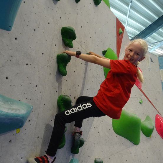 Ein junges Mädchen in einem roten Shirt klettert an einer Kletterwand mit grünen Griffen. Sie lächelt und scheint sich zu amüsieren.