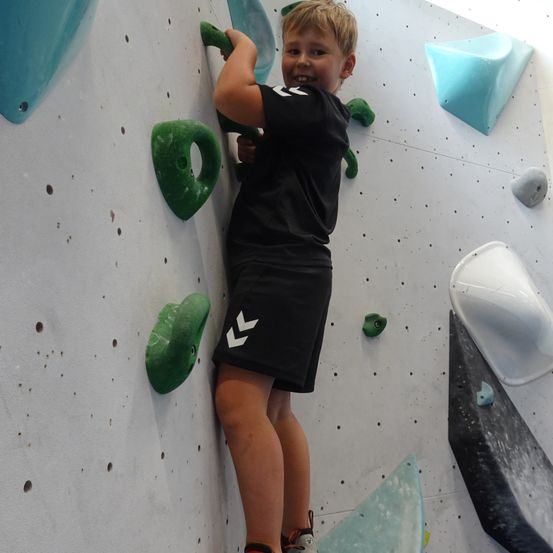 Ein junger Junge klettert an einer Indoor-Kletterwand mit verschiedenen Griffen und Haltegriffen an der Wand. Er trägt ein schwarzes T-Shirt und Shorts mit einem weißen Logo.