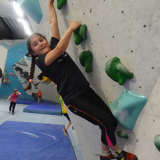 Ein Mädchen klettert in einem Fitnessstudio an einer Kletterwand. Sie trägt ein schwarzes Shirt und Hose. Im Hintergrund sind andere Leute zu sehen.
