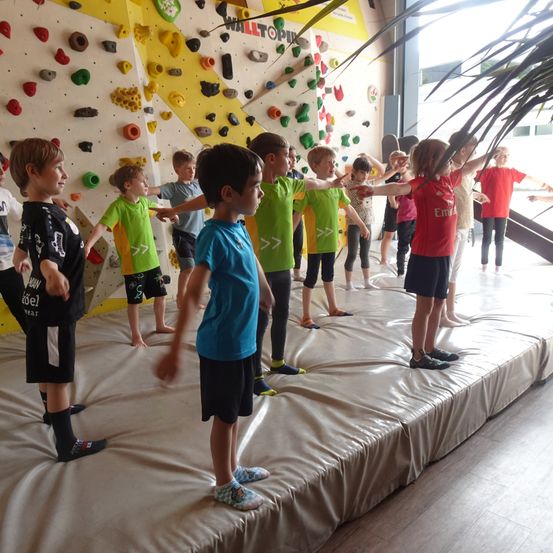 Eine Gruppe von Kindern in verschiedenen farbigen T-Shirts steht in einer Reihe auf einer gepolsterten Matte vor einer Kletterwand.