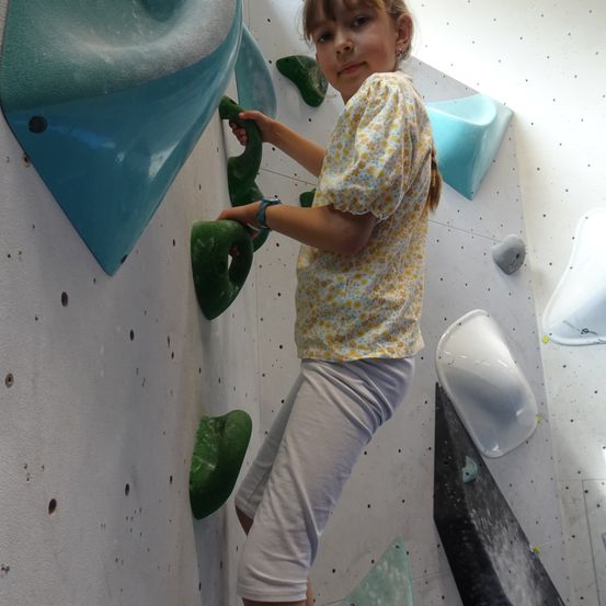 Ein junges Mädchen klettert an einer Wand mit grünen und blauen Griffen. Sie trägt ein gelbes Shirt mit weißen Hosen.
