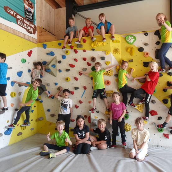 Eine Gruppe von Kindern in einem Kletterzentrum, einige klettern an der Wand, während andere sitzen oder stehen. Die Wand ist mit Klettergriffen bedeckt.