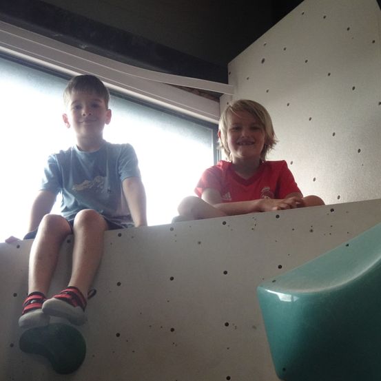 Zwei Jungen sitzen auf einer Kletterwand, einer in einem roten T-Shirt, der andere in einem blauen. Sie lächeln und schauen in die Kamera, mit einem grünen Stuhl in der Nähe.