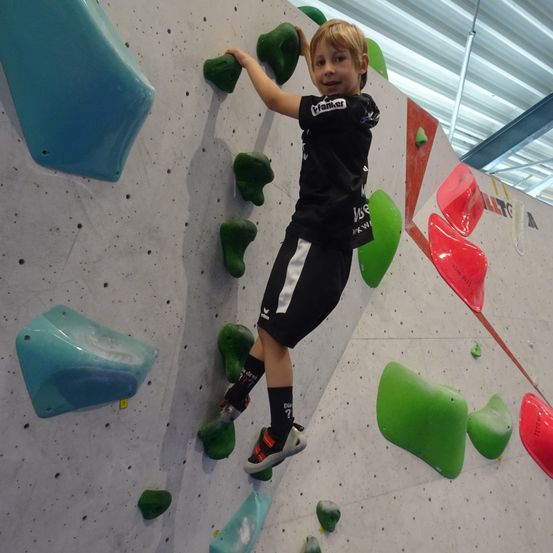 Ein junger Junge klettert an einer Kletterwand. Er trägt ein schwarzes Shirt und Shorts, dazu passende Turnschuhe. Verschiedene farbige Griffe sind an der Wand befestigt.
