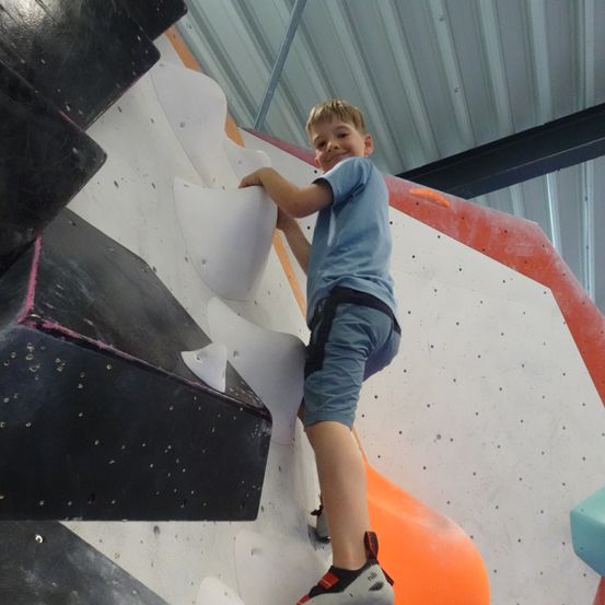 Ein junger Junge klettert eine Kletterwand, wobei seine rechte Hand an der Wand und sein linkes Bein auf einem Tritt ist. Er trägt ein blaues T-Shirt, Shorts und Turnschuhe. Die Wand ist weiß gestrichen mit orangen und schwarzen Akzenten.